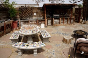 Eine Terrasse im Freien mit mozaikbesetzten Tischen und Bänken, einem Grill und einem Holzzaun.
