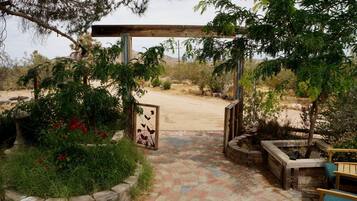 A garden entrance with a wooden pergola, a metal gate, and a paved pathway leading to a sandy area.
