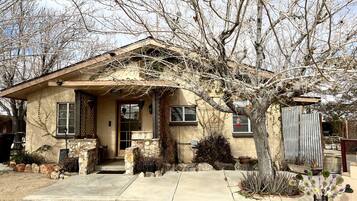 A single-story house with a sloped roof, a front porch, and a small front yard with plants and a tree.