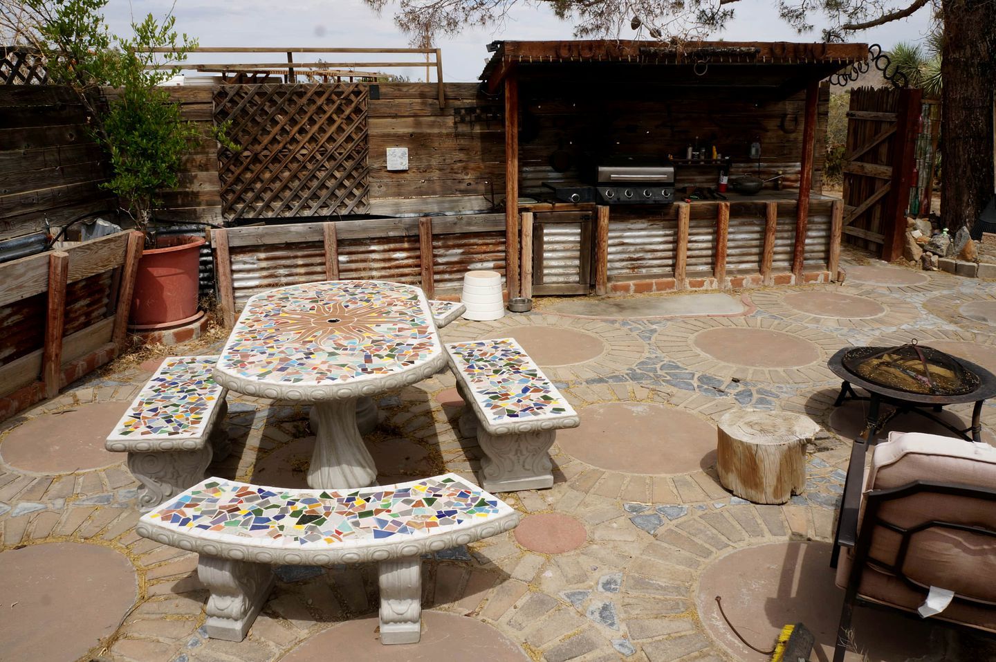 An outdoor patio with mosaic-tiled tables and benches, a barbecue grill, and a wooden fence.