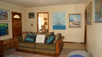 A living room with a sofa, a wooden side table, and a chair. There are paintings on the walls and a door with a window.