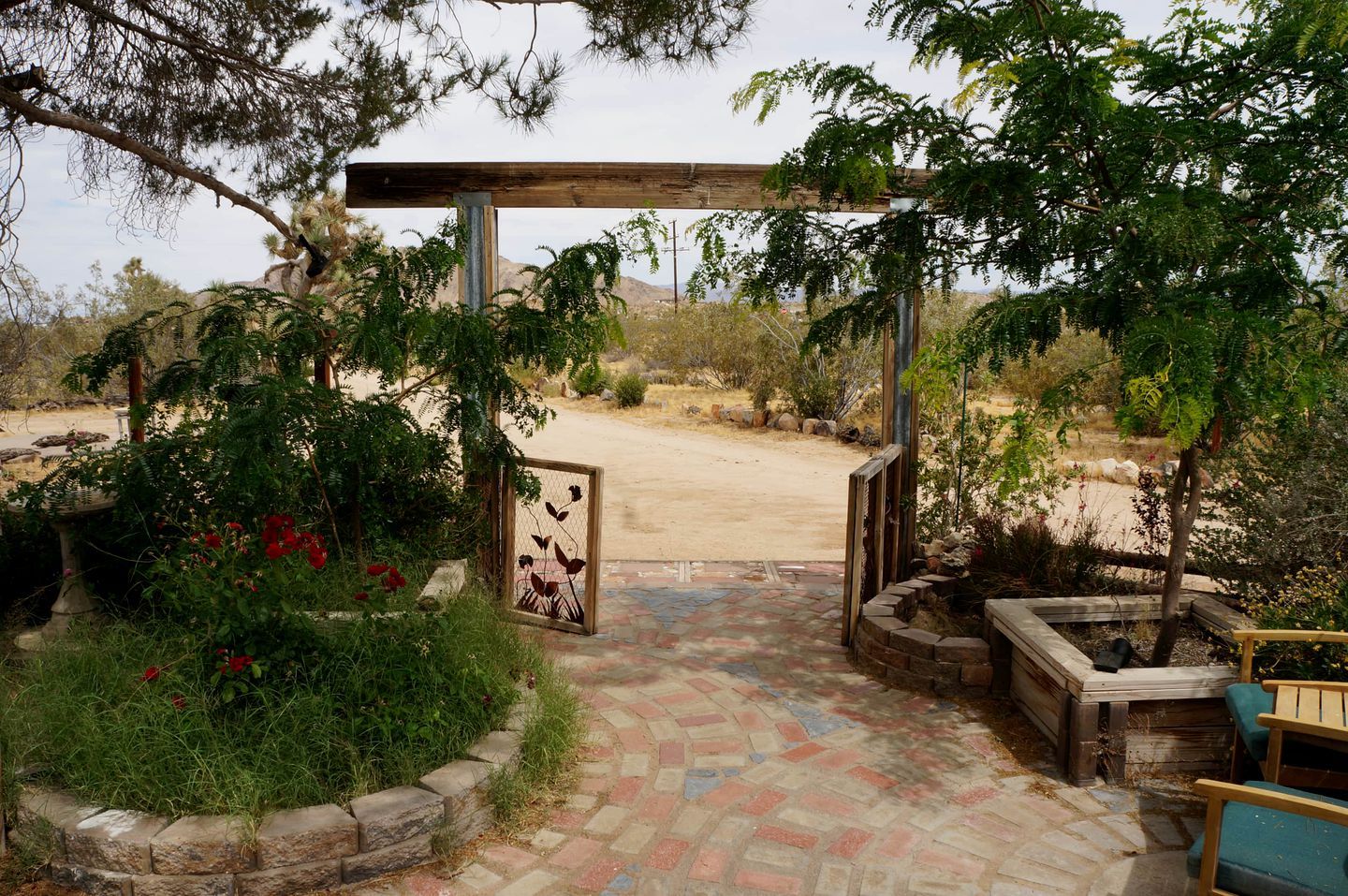 Una entrada al jardín con un pérgola de madera, un portón de metal y un camino pavimentado que conduce a una zona arenosa.