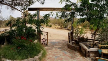 Una entrada al jardín con un pérgola de madera, un portón de metal y un camino pavimentado que conduce a una zona arenosa.