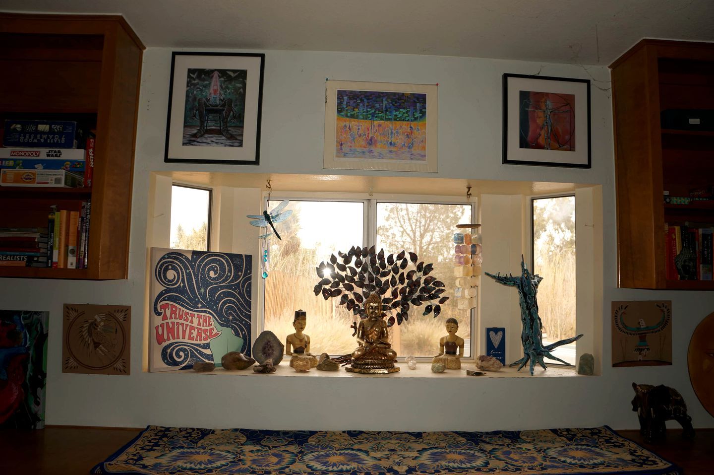 A room with a large window, a Buddha statue, a tree sculpture, and various framed artworks.