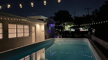 Une piscine avec des guirlandes lumineuses au-dessus, un bâtiment doté de fenêtres et un arbre en arrière-plan.