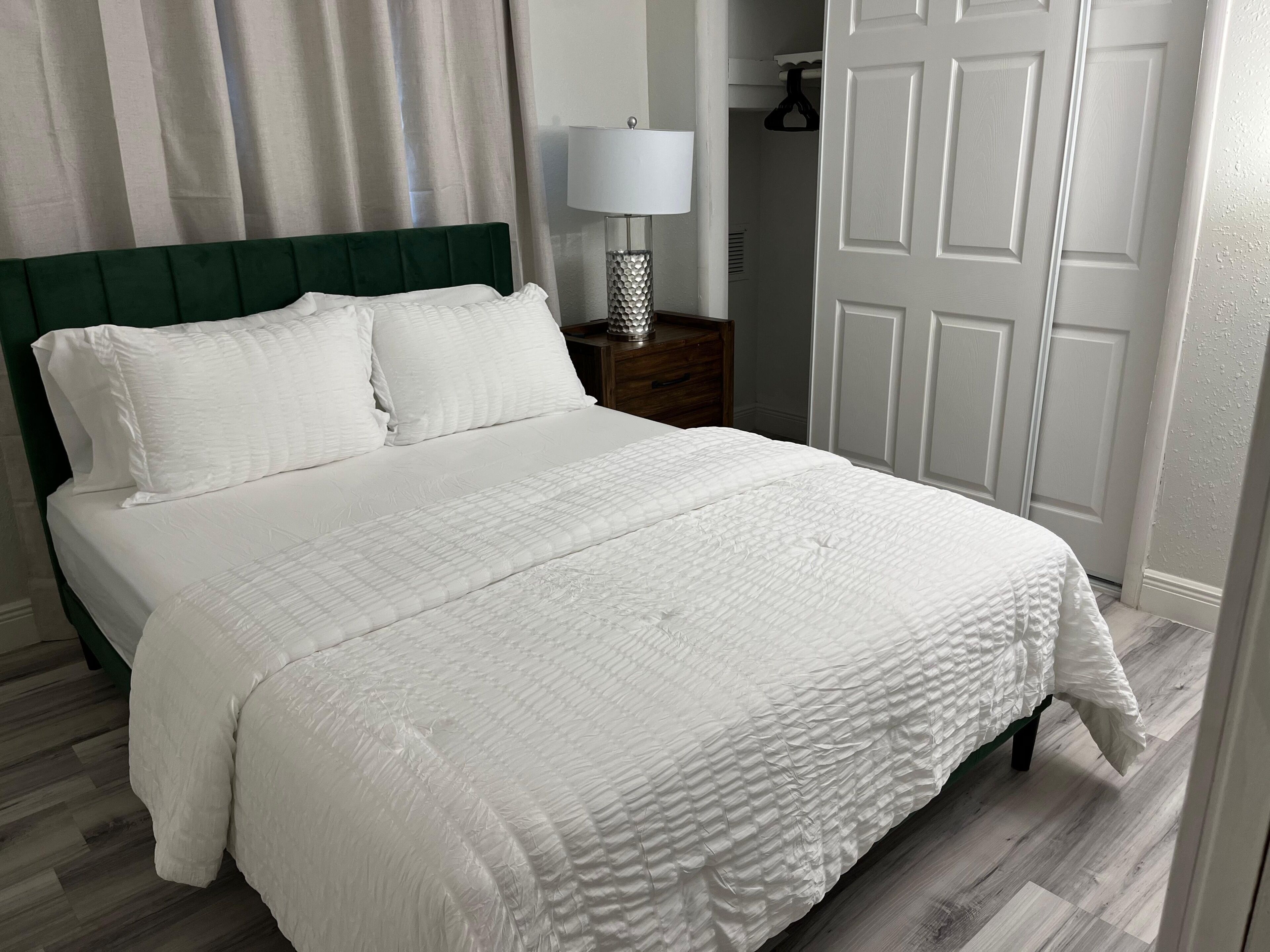 A neatly made bed with white bedding, a green upholstered headboard, and a bedside lamp on a wooden nightstand.