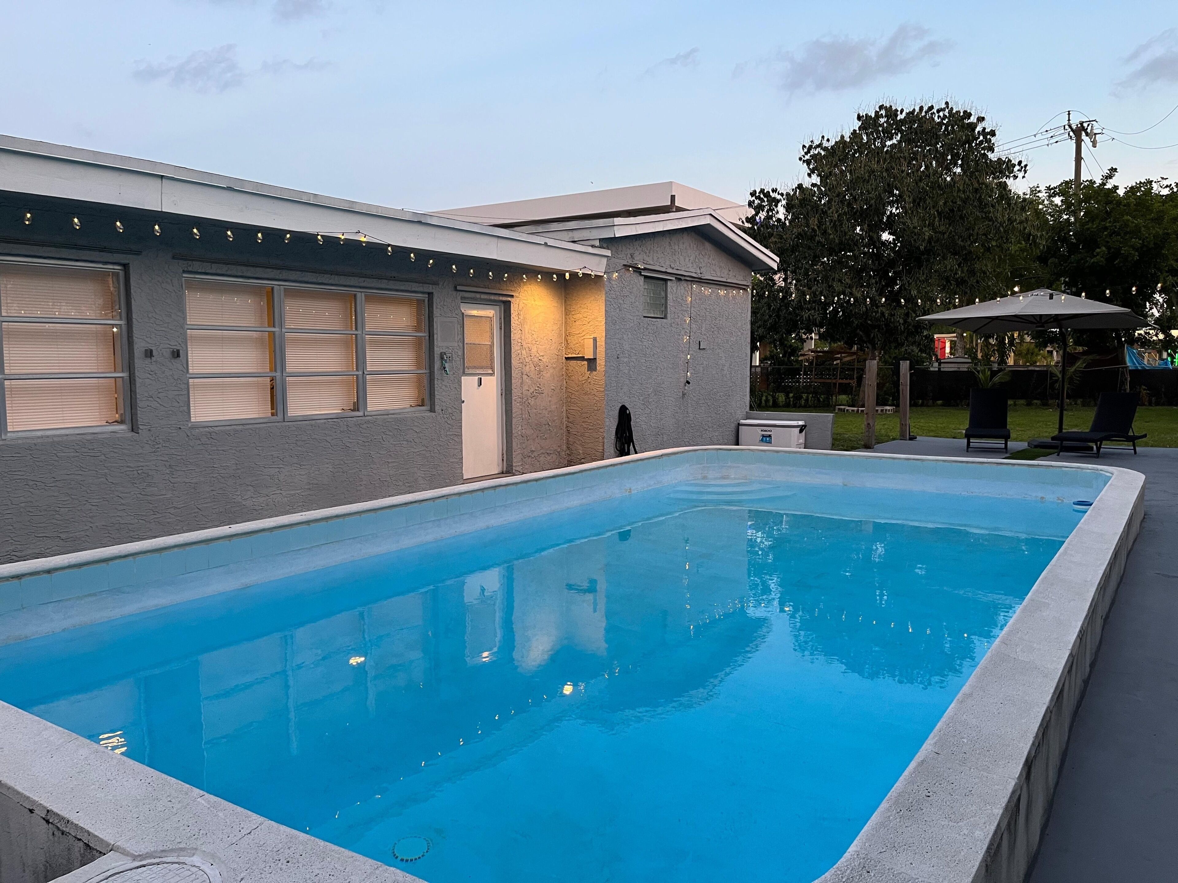 Una piscina rectangular junto a un edificio con amplios ventanales y puerta, un área de estar cubierta y una zona ajardinada con árboles.