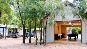 A covered outdoor area with a pool table, surrounded by trees and a building with a sloped roof. - Luxury Tented Cabin Retreat - Fun Glamping Vacation by the Water for the Whole Family in Seguin, Texas (Seguin)