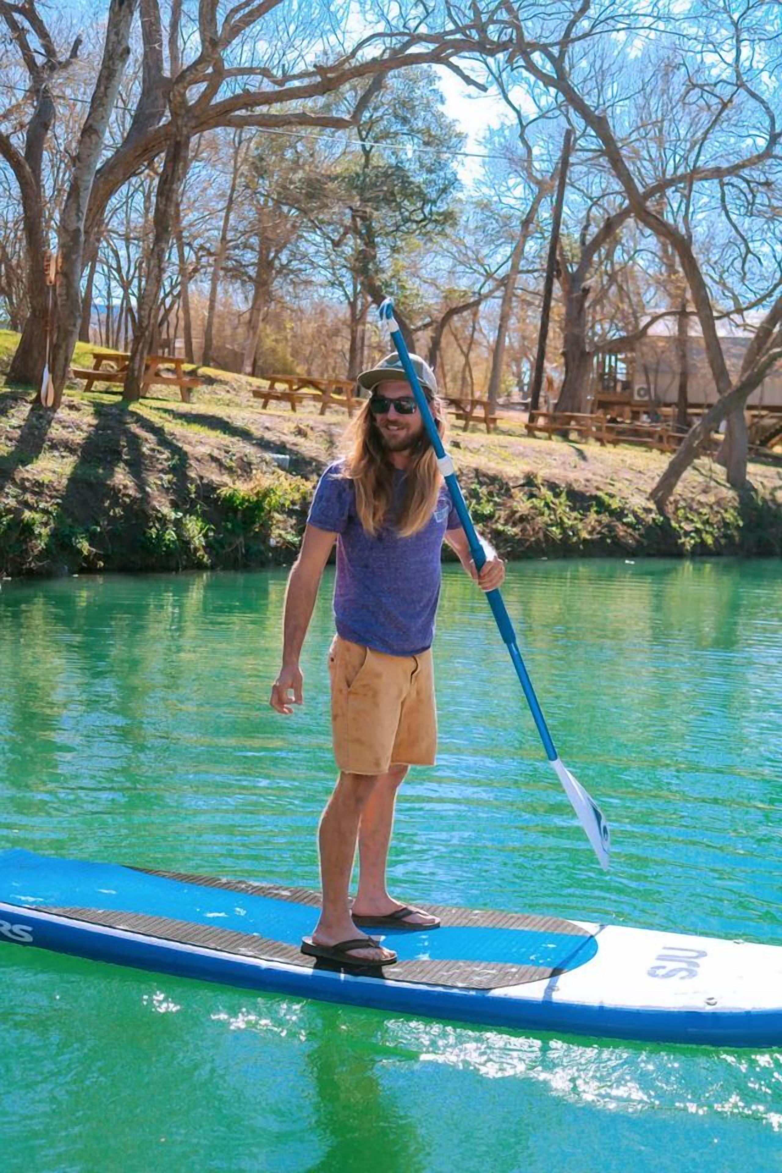 Une personne se tient debout sur une planche de paddle, sur une étendue d’eau, avec des arbres en arrière-plan.