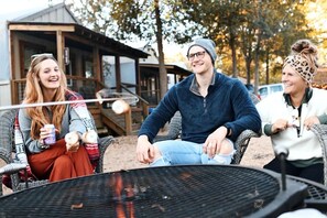 Three people sitting around a campfire in an outdoor setting with a wooden structure in the background.