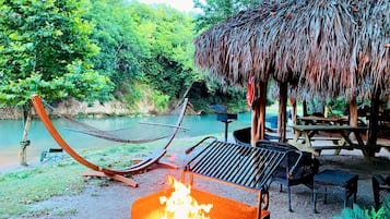 A hammock, a grill, and a thatched-roof structure by a river.