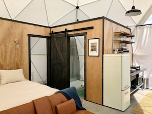 A bedroom with a bed, a dresser, a refrigerator, and a sliding door leading to a shower.