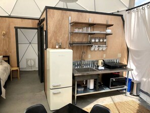 A compact kitchenette with a refrigerator, sink, and shelves for dishes.