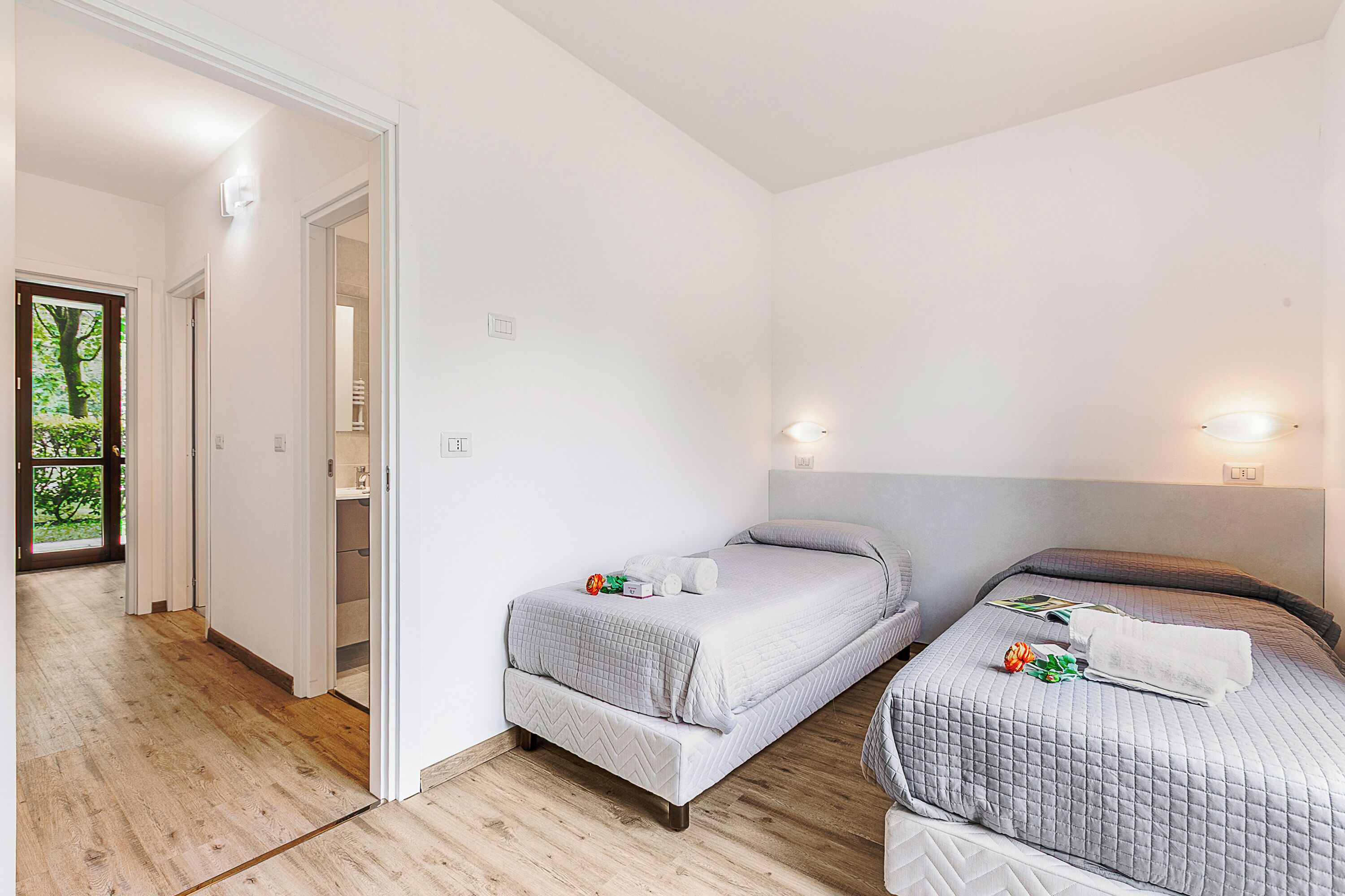 A hotel room with two single beds, a wooden floor, white walls, and a view through an open door.