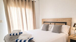 A neatly made bed with pillows, a bedside table with a lamp, and a window with curtains. - Orihuela Costa Relax & Sea (Alicante)