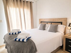 Een netjes opgemaakt bed met kussens, een nachtkastje met een lamp, en een raam met gordijnen.