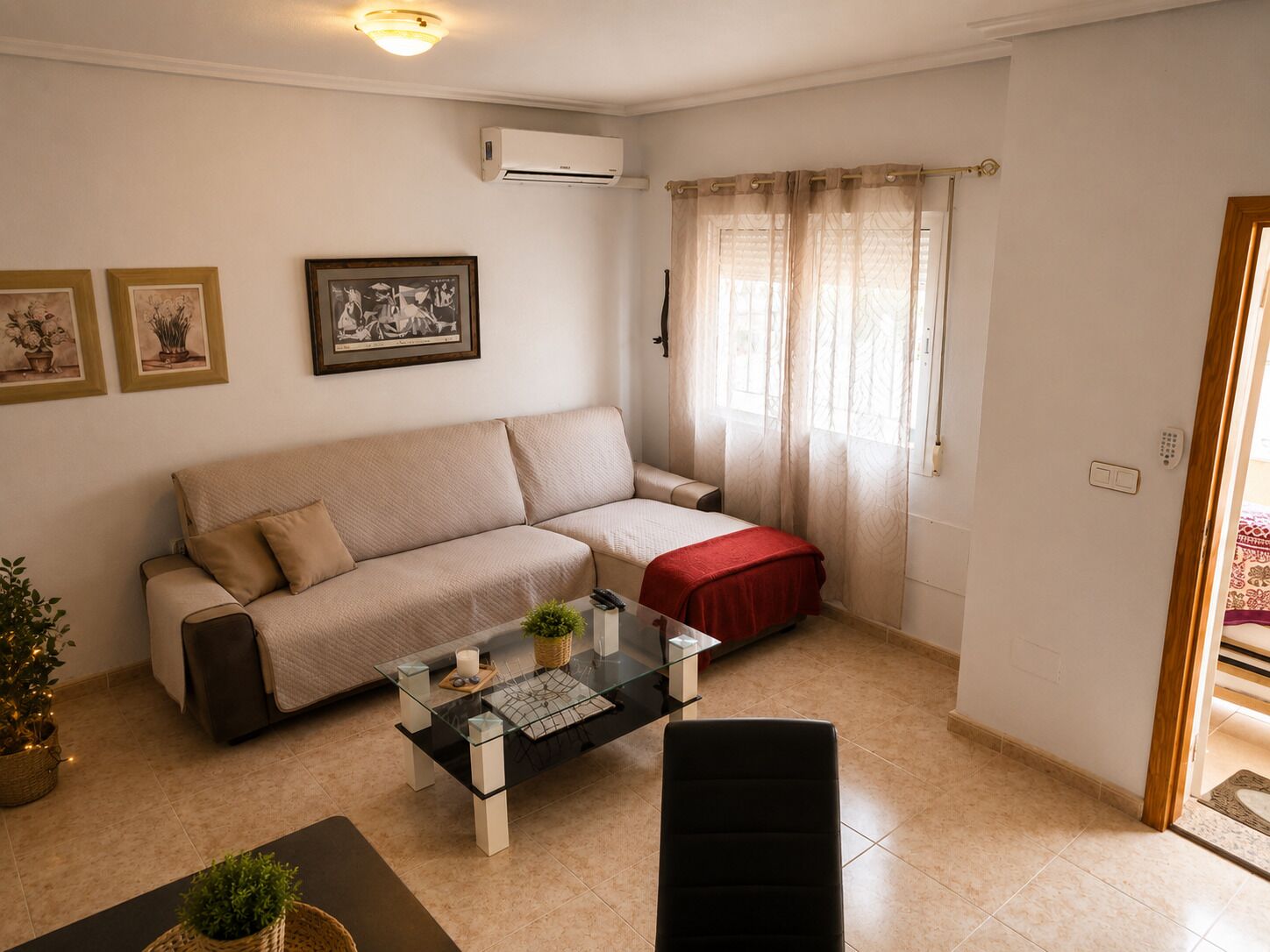 Una sala de estar con un sofá, una mesita de centro de vidrio y una pequeña silla negra. Hay una ventana con cortinas translúcidas y un aire acondicionado montado en la pared.