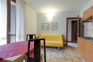 Ein kleines Zimmer mit einem Bett, einem kleinen Tisch, einem Sessel, einer Küchenzeile und zwei gerahmten Bildern an der Wand.
