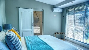 Un dormitorio con cama, mesita de noche, espejo, armario, ventana con persianas y unidad de aire acondicionado.