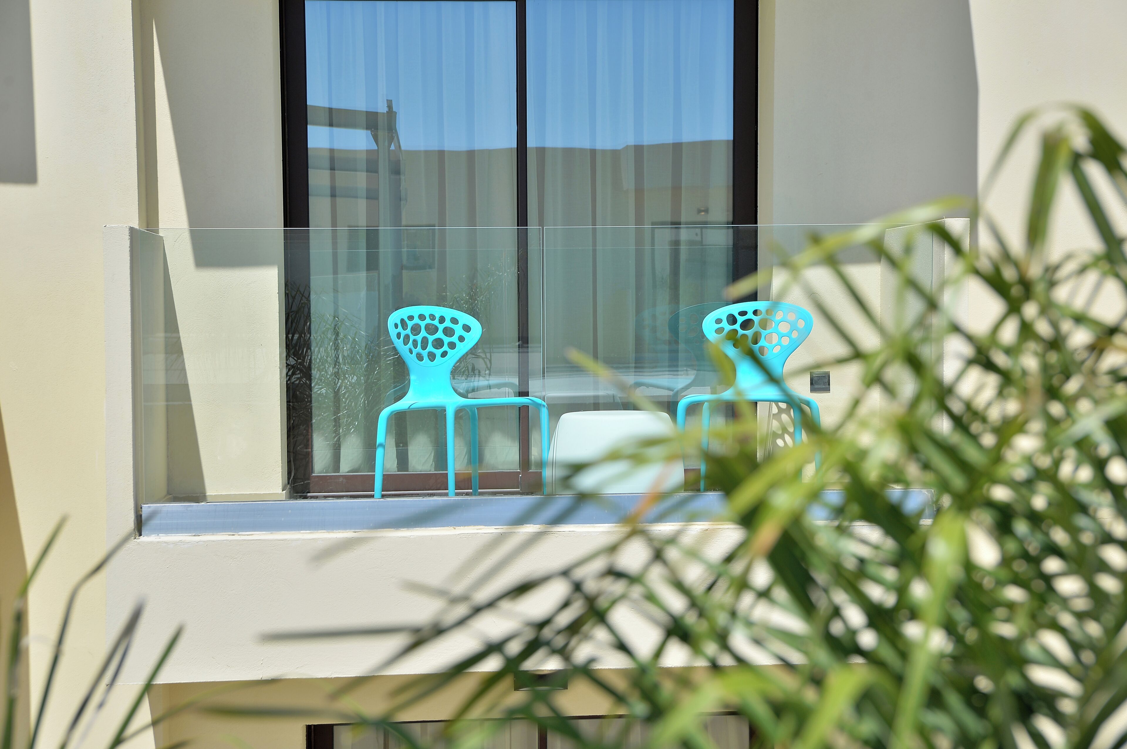 A balcony with a glass railing, two turquoise chairs, and a table.