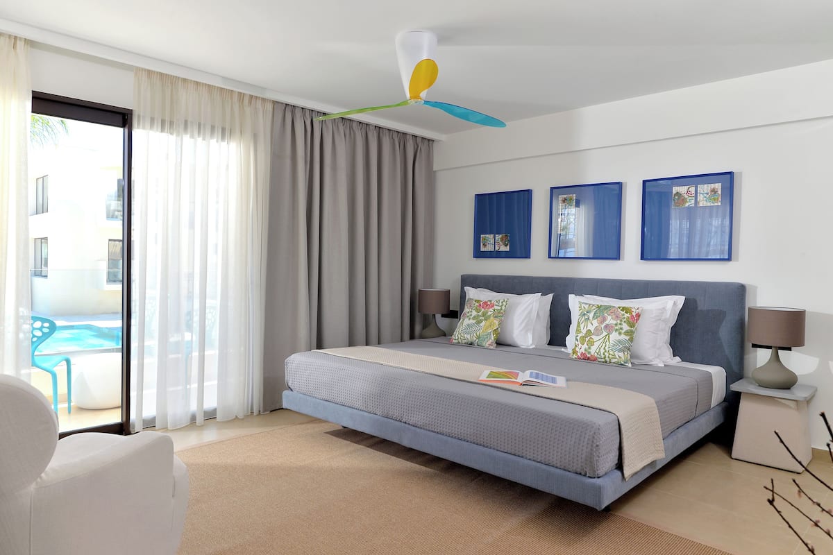 Habitación de hotel con una cama, un ventilador de techo, vista a una piscina y dos cuadros en la pared.