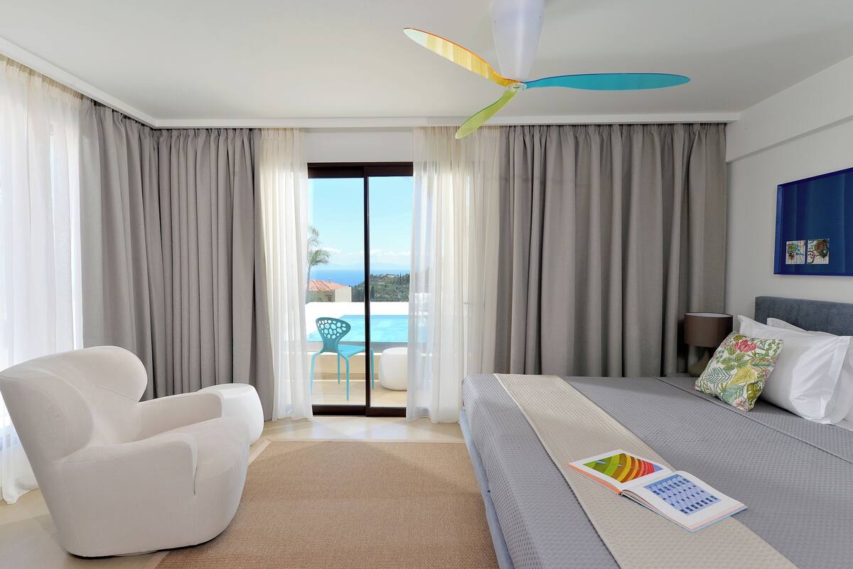 Una habitación de hotel moderna con una cama, un sillón blanco, un ventilador de techo y vistas a una piscina.