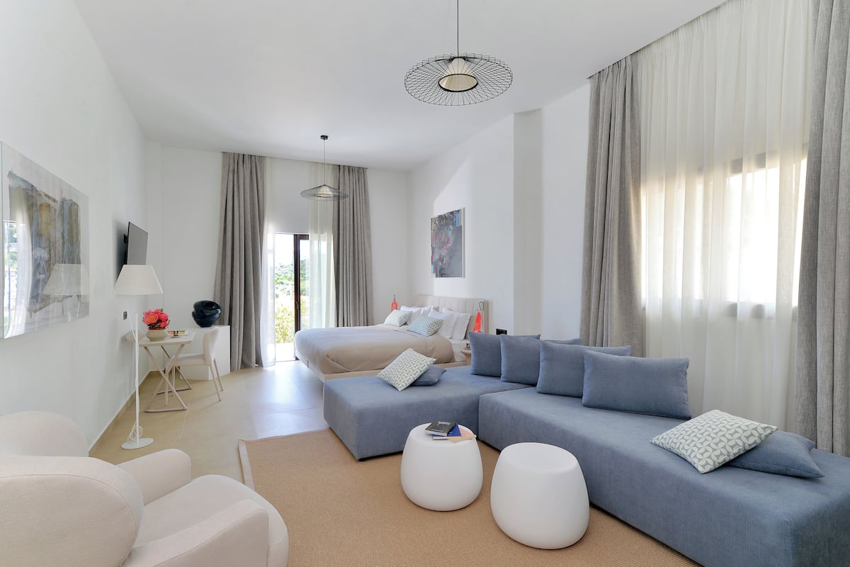 Una habitación de hotel moderna con una cama grande, un sofá gris, una mesita pequeña y un sillón blanco.