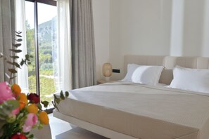 A bedroom with a large bed, a bedside table with a lamp, and a view of greenery through the window. - Althea Resorts (Xylokastro-Evrostina)