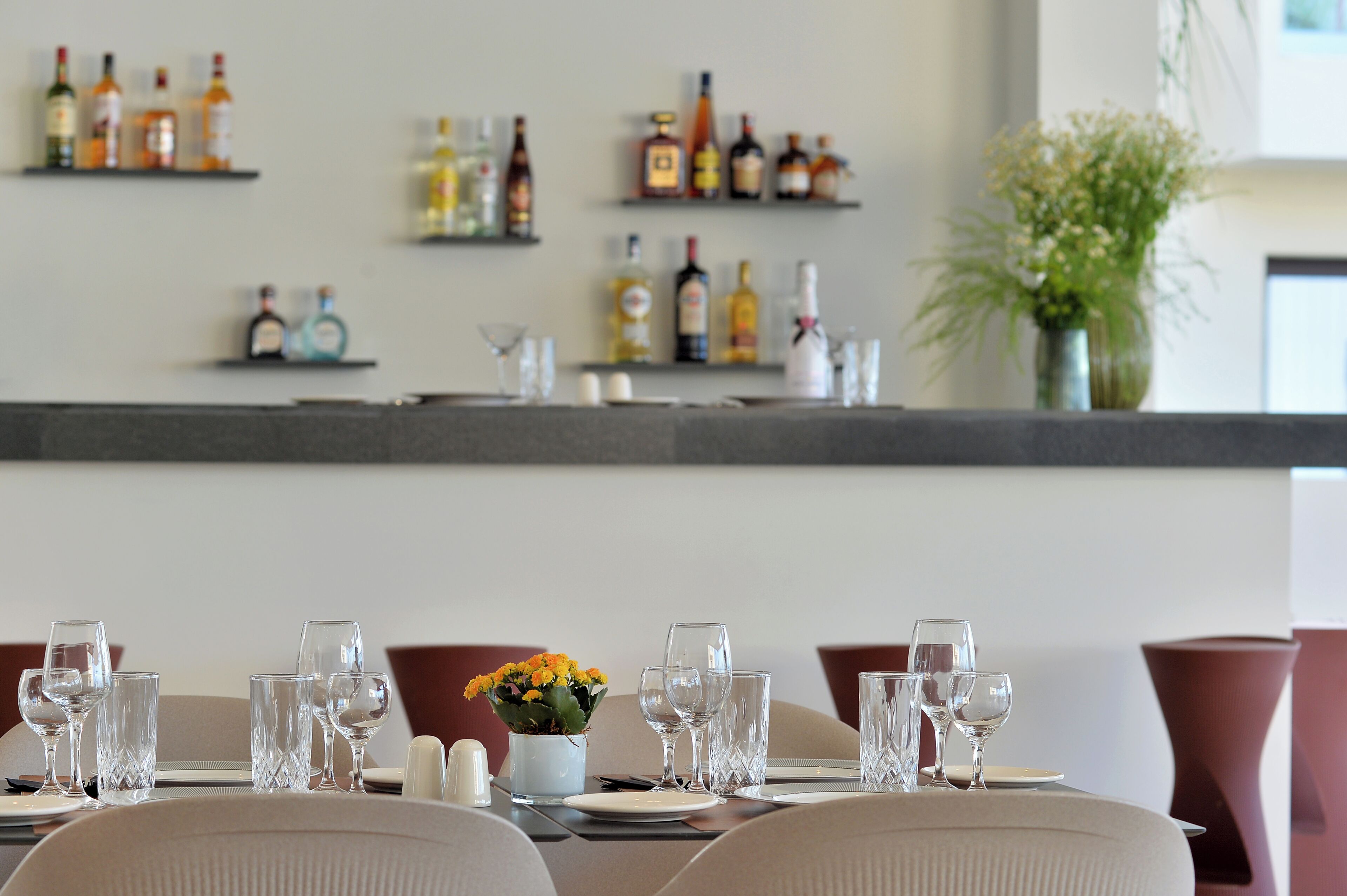 A dining table set with glasses and a vase of flowers, with shelves of bottles in the background.