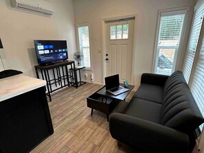 Ein Wohnzimmer mit einer Couch, einem Couchtisch mit einem Laptop, einem Fernseher und einem Ventilator.