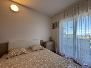 A bedroom with a bed, a nightstand, a dresser, a window with curtains, and a wall-mounted light.