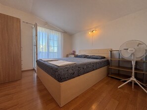 A bedroom with a bed, a standing fan, a window with curtains, and a door.