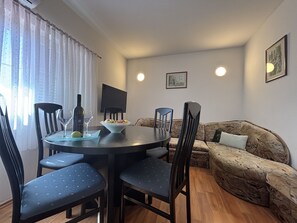 A dining area with a round table, chairs, a bowl of fruit, a wine bottle, and a glass. There is a sofa and a television in the background.