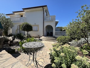 A two-story house with a balcony, a stone patio, and a garden with various plants.