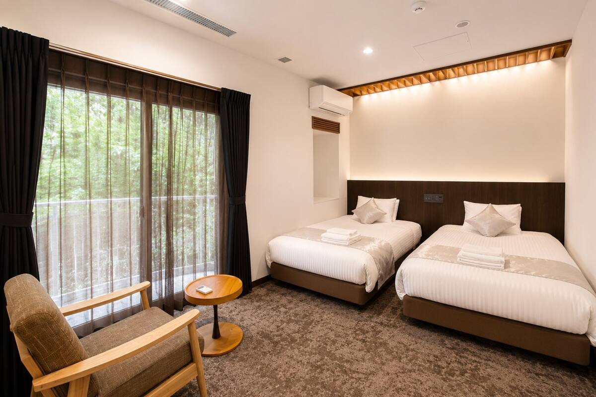 Habitación de hotel con dos camas, una silla, una mesita pequeña y un ventanal grande con cortinas.