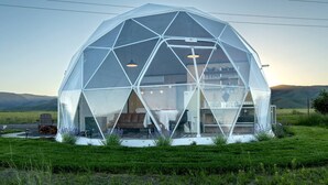 A geodesic dome structure with a visible interior including a table and chairs, surrounded by a grassy field and hills in the background.