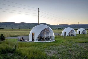 Trois structures en forme de dôme géodésique, situées dans un cadre rural, avec un ciel dégagé et des montagnes en arrière-plan.