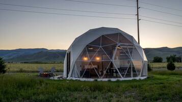 A geodesic dome structure with a visible interior, surrounded by a grassy field and hills in the background.