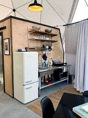 Une cuisine de taille réduite avec un réfrigérateur blanc, un évier et une cuisinière. Des étagères au-dessus du plan de travail servent à ranger la vaisselle et les verres. On aperçoit une chaise noire au premier plan.