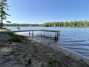 Eine Seenlandschaft mit einem hölzernen Steg, der sich ins Wasser erstreckt, umgeben von Bäumen und wolkenlosem Himmel.