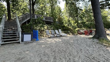 Zona de playa arenosa con una escalera de madera que lleva a una terraza, rodeada de árboles.