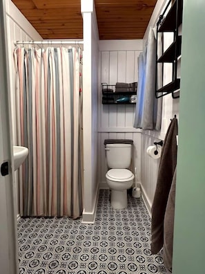 A small, compact bathroom with a striped shower curtain, a toilet, a shelf with towels, and patterned floor tiles. - Cozy Vintage Lake Cabin with Hot Tub & Beach Access near Traverse City, Northern Michigan (Traverse City)