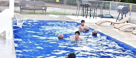 Una piscina con aro de baloncesto, sillas de descanso y un área cercada con árboles.