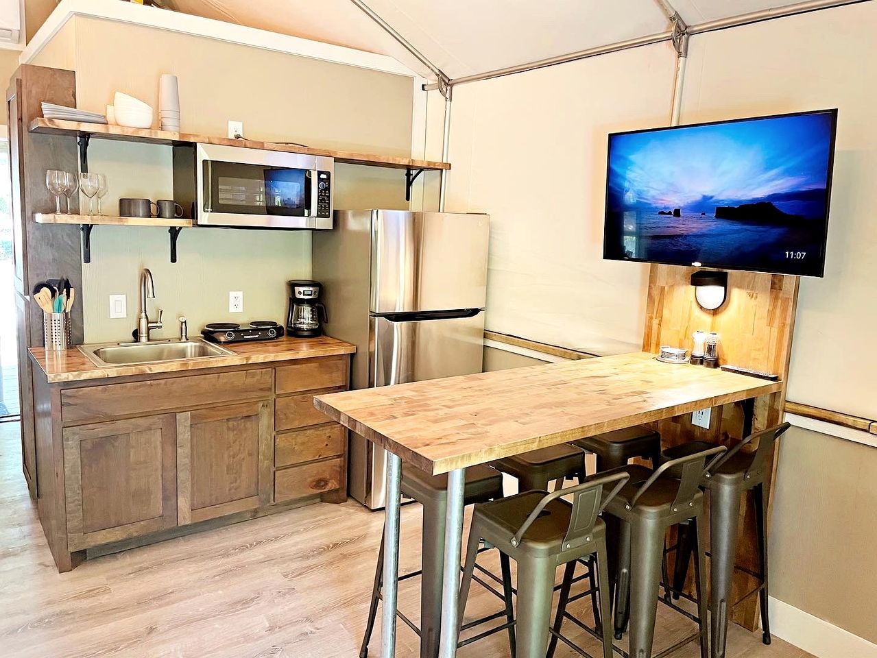 Ett kompakt kök med ett träbord, barstolar, en mikrovågsugn, ett kylskåp och en platt-TV som är monterad på väggen.