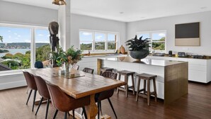 Een moderne keuken met een groot eiland, een houten eettafel en stoelen, grote ramen met uitzicht en een hanglamp.