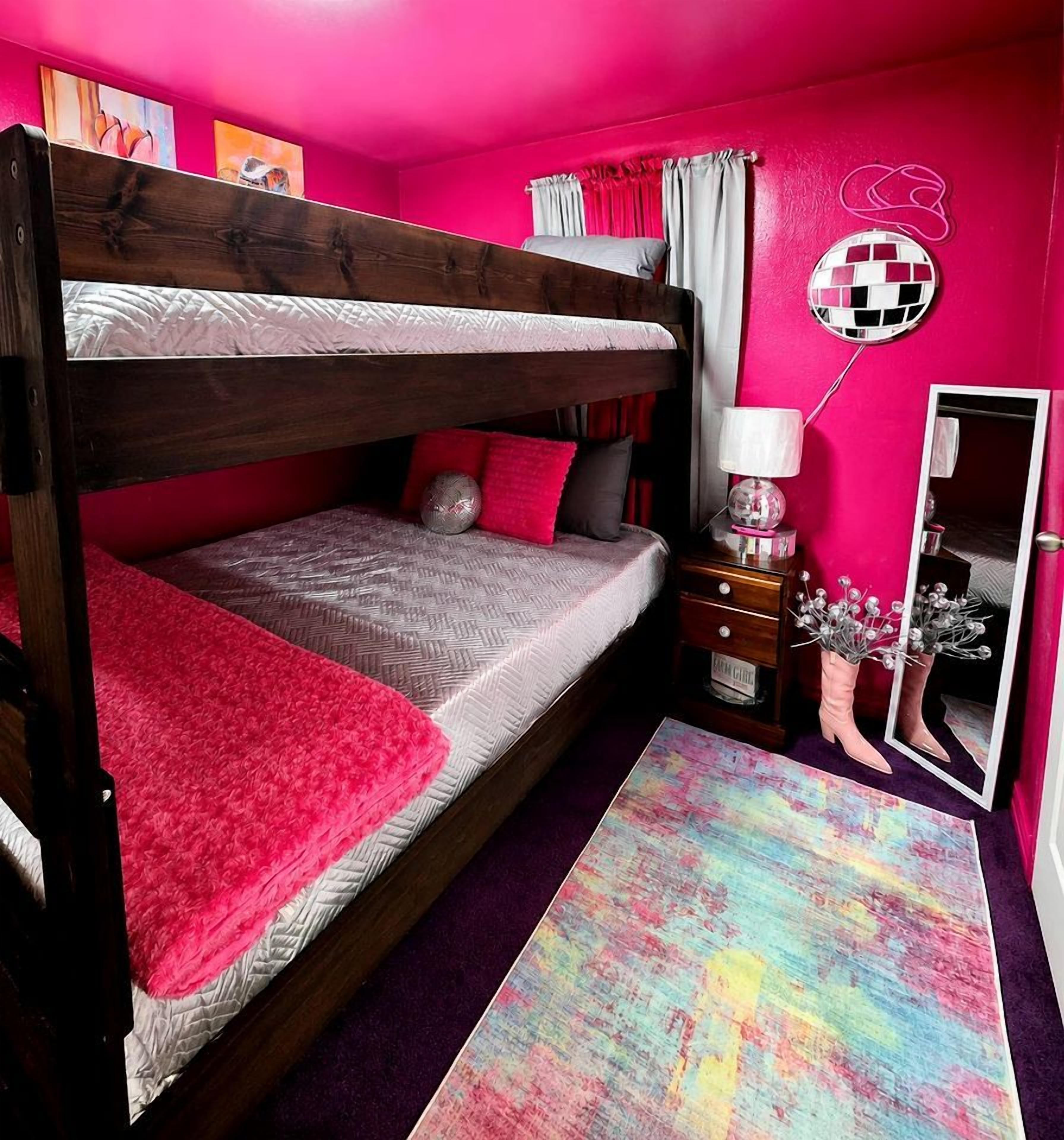 Ein Etagenbett mit roter Decke, ein Spiegel, eine Kommode und ein farbenfroher Teppich in einem Zimmer mit rosafarbenen Wänden.