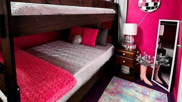 Ein Etagenbett mit roter Decke, ein Spiegel, eine Kommode und ein farbenfroher Teppich in einem Zimmer mit rosafarbenen Wänden.