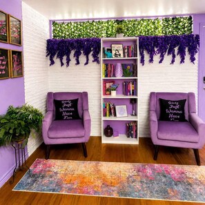 Ein Zimmer mit zwei violetten Sesseln, einem Bücherregal und einer weißen Ziegelwand.