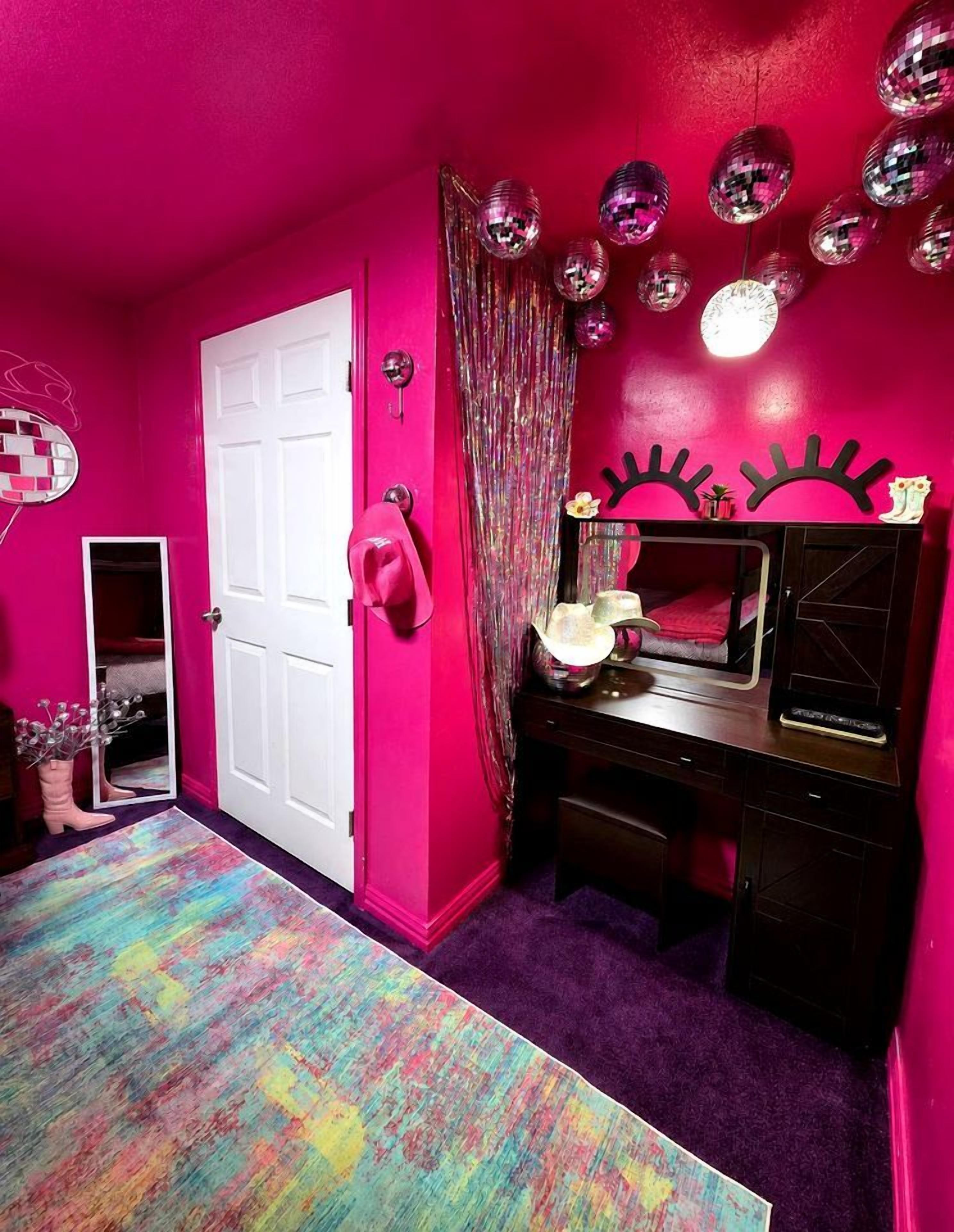 Una habitación con pared rosa, puerta blanca, espejo, tocador con lámpara y una alfombra colorida.