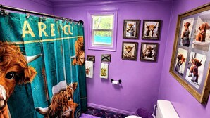 A bathroom with a purple wall, a shower curtain with a cow design, and several framed pictures on the wall.